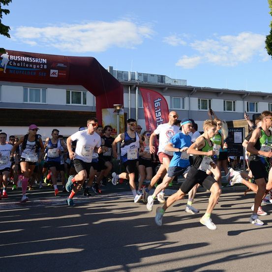 Teilnehmer eines Marathonlaufs, die unter einem Banner mit der Aufschrift 'BUSINESSRUN Challenge2B' laufen. Die Läufer tragen nummerierte Startnummern und Turnschuhe. Im Hintergrund ist ein Gebäude mit einem roten Banner zu sehen.