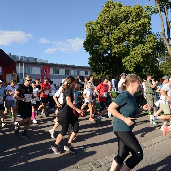 Eine Gruppe von Läufern nimmt an einem Marathon teil, während Zuschauer sie von den Seitenlinien anfeuern. Die Läufer tragen verschiedene farbige Shirts und Turnschuhe. Ein Gebäude mit einem roten Banner ist im Hintergrund zu sehen.