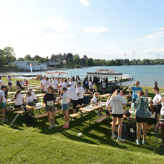 Eine Gruppe von Menschen in weißen Shirts und Shorts versammelt sich auf einem Rasenfeld in der Nähe eines Sees, einige sitzen an Picknicktischen, andere stehen. Eine Bar mit einem Schild, das 'BIER-BAR' steht auf der Anlegestelle. Bäume und Gebäude sind im Hintergrund zu sehen.