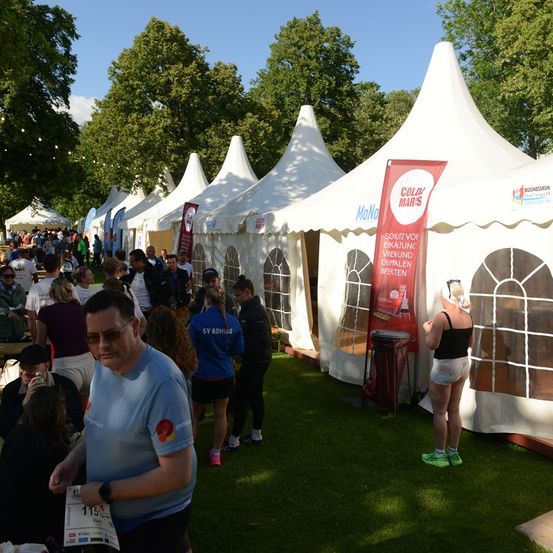 Eine große Gruppe von Menschen versammelt sich in einem Park mit mehreren aufgestellten Zelten. Einige tragen Sportausrüstung und halten Nummern, was auf eine Marathonveranstaltung hindeutet. Bäume und ein klarer Himmel sind im Hintergrund sichtbar.
