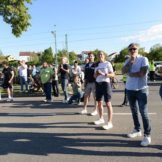 Eine Gruppe von Menschen steht auf einer Straße, einige machen Fotos, während ein Mann in einem weißen Shirt in ein Mikrofon spricht. Im Hintergrund sind Bäume und Häuser zu sehen.