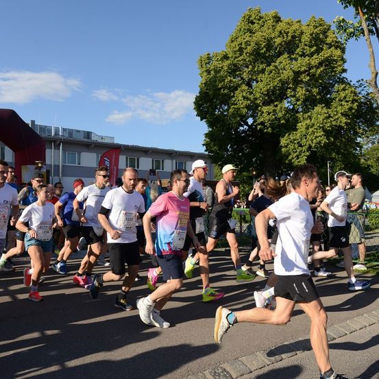Eine Gruppe von Läufern bei einem Marathon, einige in weißen Shirts und andere in bunten Klamotten, laufen auf einer Straße mit Bäumen und Gebäuden im Hintergrund.