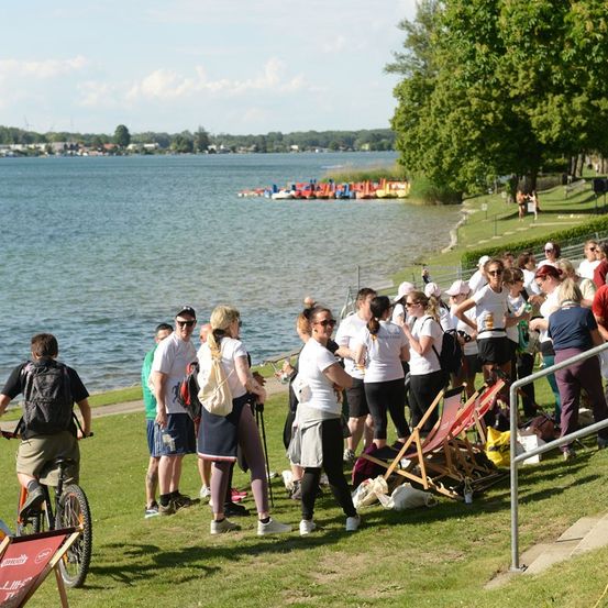 Eine Gruppe von Menschen in weißen Shirts versammelt sich in der Nähe eines Sees, einige trinken und andere gehen. Es gibt Boote im Wasser und Bäume im Hintergrund.