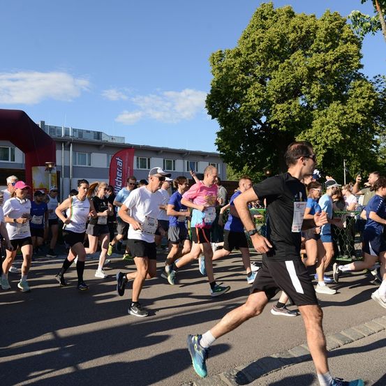 Eine Gruppe von Läufern nimmt an einem Marathon teil, wobei ein Mann in Schwarz die Gruppe anführt, gefolgt von anderen in verschiedenen Farben. Das Rennen findet an einem sonnigen Tag mit klarem blauem Himmel statt.