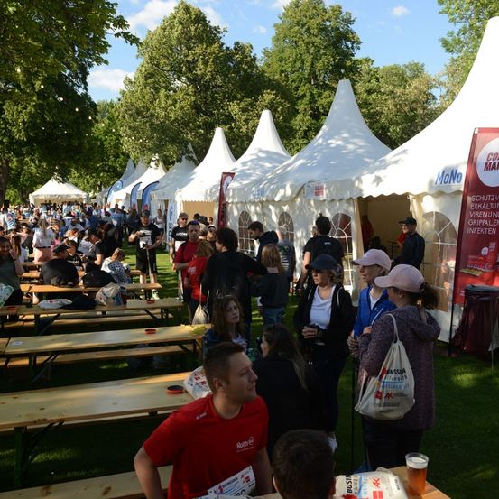 Eine große Menschenmenge versammelt sich unter weißen Zelten in einem Park, einige in roten Shirts, andere in Freizeitkleidung. Tische und Bänke sind aufgestellt, und die Leute gehen herum, unterhalten sich und genießen die Veranstaltung im Freien.