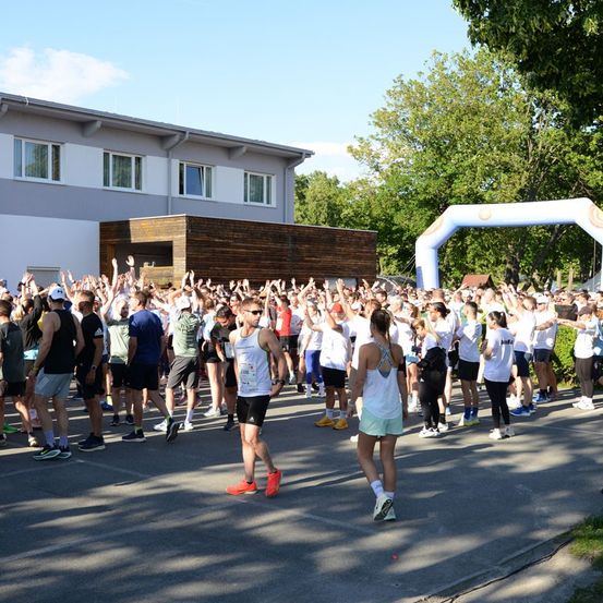 Eine große Gruppe von Menschen in Laufbekleidung versammelt sich vor einem Gebäude, einige heben ihre Hände. Der Ort ist im Freien mit Bäumen und einem klaren Himmel.