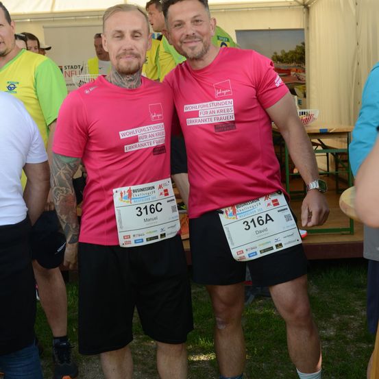 Zwei Männer in rosa T-Shirts mit der Aufschrift Wohlfuhlstunden fur an Krebs Erkrankte Frauen stehen zusammen vor einer Menschenmenge. Sie tragen beide schwarze Shorts und haben Nummern auf ihren Shirts.