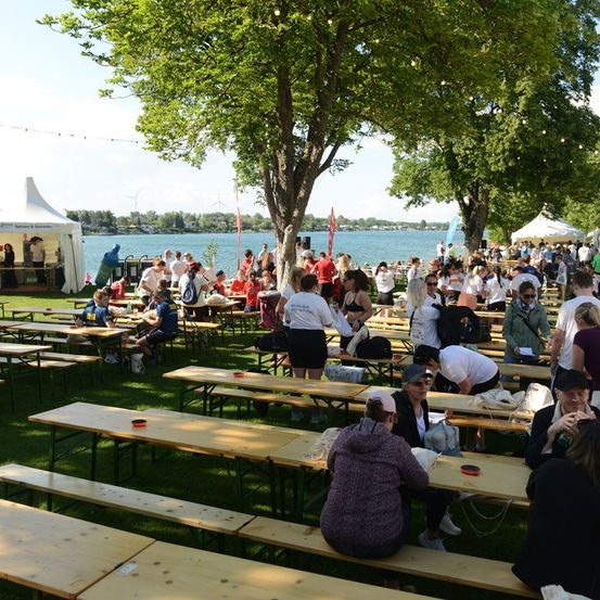 Eine große Gruppe von Menschen versammelt sich in einer Outdoor-Umgebung, möglicherweise für ein Festival. Sie sitzen an Picknicktischen auf einem Rasenfeld mit einem See und Bäumen im Hintergrund. Einige Leute tragen Hüte und Sonnenbrillen, und in der Ferne sind Zelte und eine Bühne zu sehen.