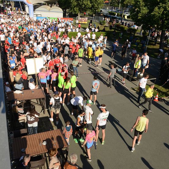 Eine große Menschenmenge in verschiedenen Laufausrüstungen versammelt sich in einem Freiluftbereich, möglicherweise für einen Marathon. Einige stehen, während andere auf Stühlen sitzen. Es gibt Tische mit Gläsern und Tellern. In der Ferne sind Bäume, ein Gebäude und ein Bus zu sehen.