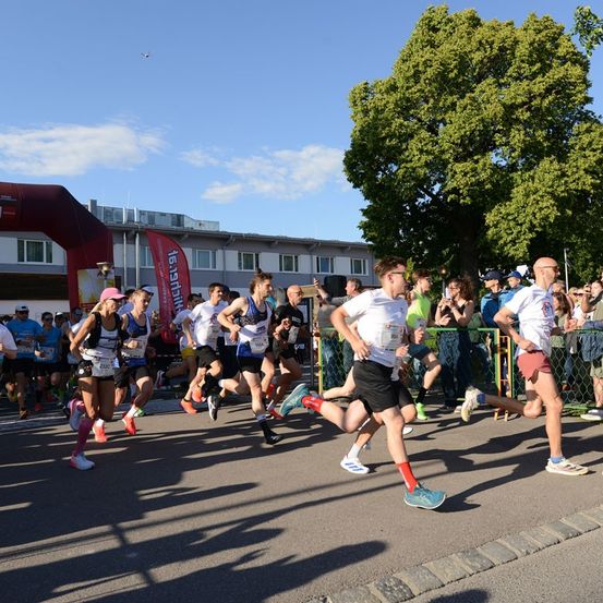 Eine Gruppe von Läufern nimmt an einem Marathon teil, während Zuschauer sie von hinter einem Zaun anfeuern. Der Himmel ist blau mit einigen Wolken.
