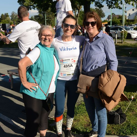 Drei Frauen stehen zusammen in einem Park und lächeln für ein Foto. Die Frau in der Mitte trägt ein weißes T-Shirt mit Viktoria und einer Startnummer. Die Frau auf der linken Seite trägt eine türkisfarbene Weste und eine Brille. Die Frau auf der rechten Seite trägt ein blau kariertes Hemd und trägt eine braune Tasche. Hinter ihnen steht ein Mann in einem weißen T-Shirt in der Nähe eines Tisches.
