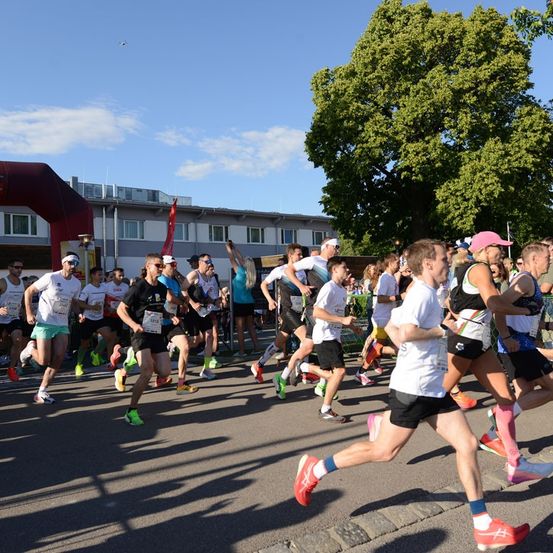 Eine Gruppe von Läufern nimmt an einem Marathon teil, der auf einer asphaltierten Straße unter einem blauen Himmel mit verstreuten Wolken stattfindet. Bäume und Gebäude säumen die Seiten der Straße.