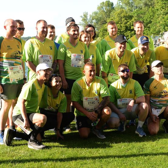 Eine Gruppe von Läufern in grünen und gelben Shirts posiert für ein Foto auf einem Rasenfeld mit Bäumen im Hintergrund.