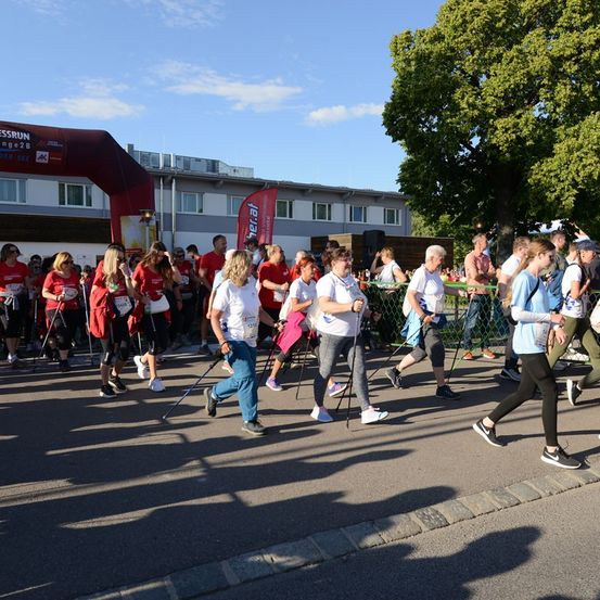 Eine Gruppe von Menschen in roten Shirts und Turnschuhen läuft in einer Reihe auf einer Straße, möglicherweise an einem Marathon teilnehmend. Im Hintergrund befinden sich Bäume und ein Gebäude.