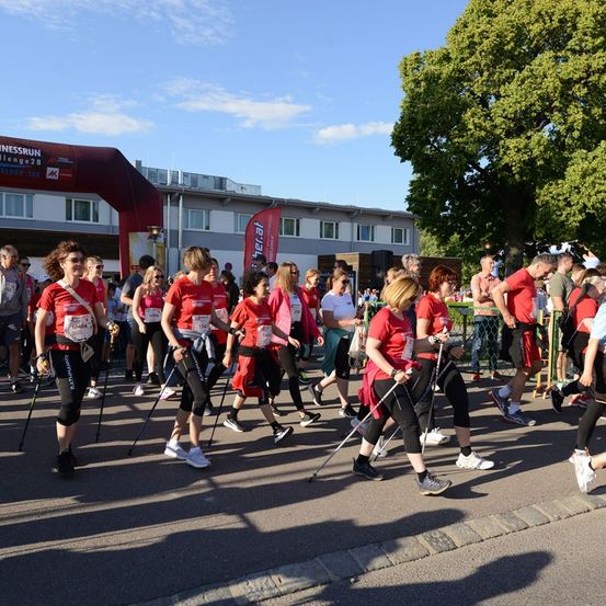 Eine Gruppe von Menschen in roten Shirts und schwarzen Hosen läuft mit Trekkingstöcken bei einem Marathon. Dahinter steht ein Gebäude mit einem Banner und einem Baum.