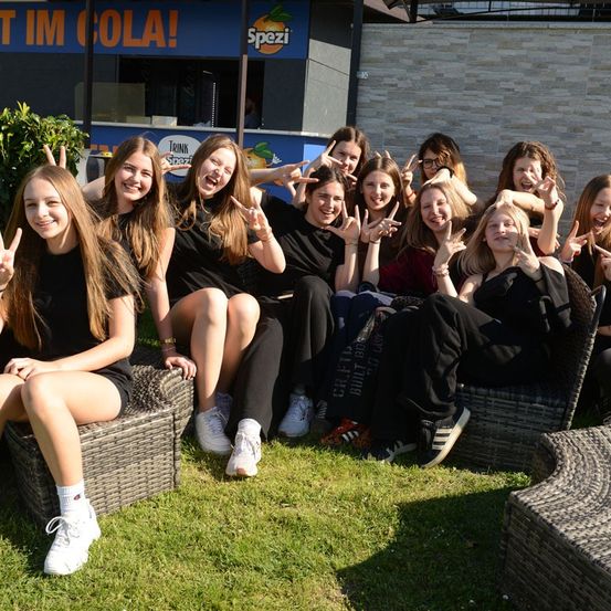 Eine Gruppe junger Frauen in passenden schwarzen Shirts sitzt auf Outdoor-Stuhlchen, lächelt und posiert für ein Foto. Sie sind vor einem Gebäude mit einem Schild, das 'Spezzi' sagt.
