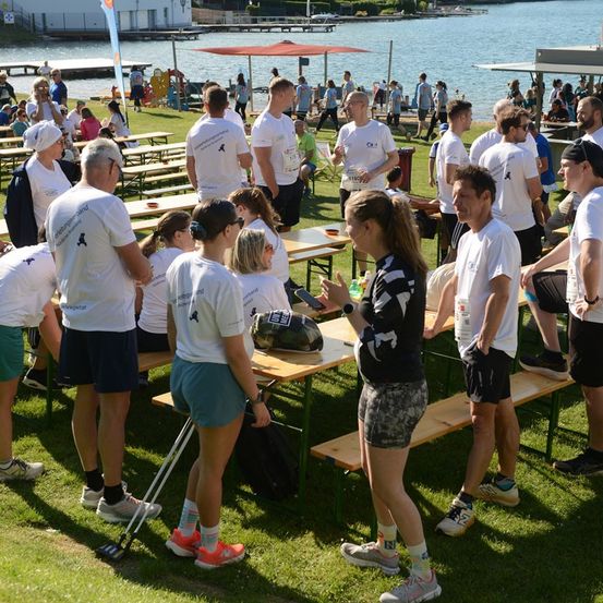 Eine Gruppe von Menschen in weißen T-Shirts versammelt sich um Tische auf einem Rasenfeld in der Nähe eines Sees, wobei einige Personen stehen und andere sitzen. Eine Frau in einem schwarzen und weißen Outfit steht vor einem Tisch und spricht möglicherweise die Gruppe an.