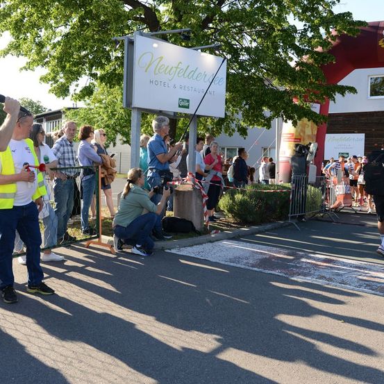 Eine Gruppe von Menschen versammelt sich um ein Schild für das Hotel und Restaurant Neufelderhof, einige machen Fotos und andere stehen daneben.