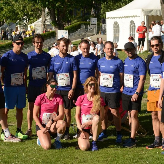 Eine Gruppe von Läufern in passenden blauen Shirts und Shorts posiert für ein Foto, wobei zwei Frauen in rosa Shirts knien. Sie tragen Startnummern und Sonnenbrillen. Im Hintergrund befinden sich Zelte und Menschen.