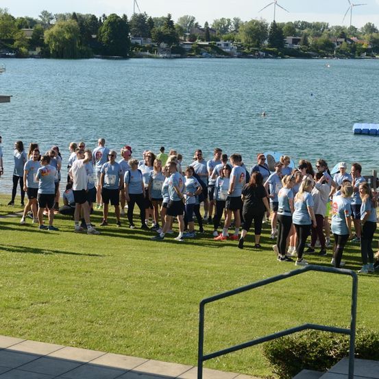 Eine Gruppe von Menschen in passenden blauen Shirts und Shorts steht auf einem Rasen neben einem See, mit klarem Himmel und Windturbinen im Hintergrund.