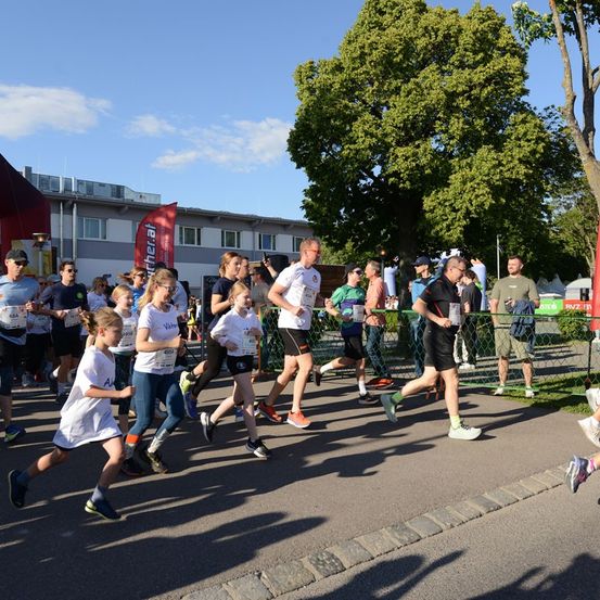 Eine Gruppe von Menschen nimmt an einem Marathon teil und läuft auf einer asphaltierten Straße, mit einem Baum und einem Gebäude im Hintergrund.