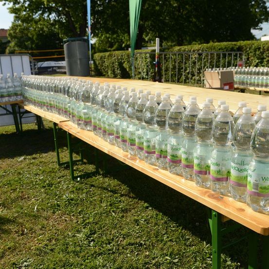 Ein langer Tisch, gefüllt mit Reihen von klaren Plastikwasserflaschen, beschriftet mit 'Waldquelle', vor einem Hintergrund aus Grün und einem Metallzaun.