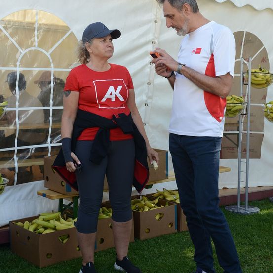 Eine Frau in einem roten AK-Shirt steht neben einem Mann in einem weißen Shirt, beide schauen auf ein Handy. Sie stehen vor einem Zelt mit Bananen im Schaufenster.