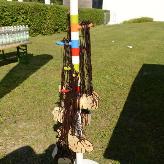 Ein farbenfroher Pfahl mit mehreren hängenden Holzanhängern auf einem Rasen, mit einem Tisch voller Wasserflaschen im Hintergrund.