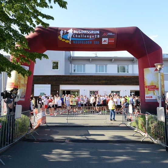 Eine Gruppe von Läufern in verschiedenen farbigen Shirts und Turnschuhen steht in einer Reihe am Start einer Veranstaltung. Der Bogen über ihnen trägt die Aufschrift 'BUSINESSRUN Challenge2B'.