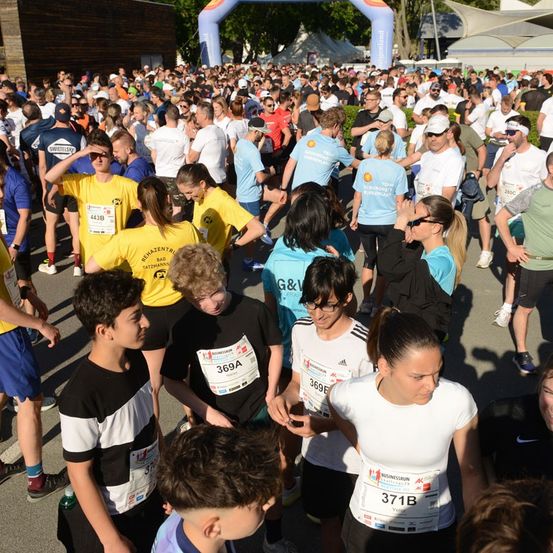 Eine große Menschenmenge mit nummerierten Startnummern versammelt sich für einen Marathon, einige gehen, während andere stehen und sich unterhalten.