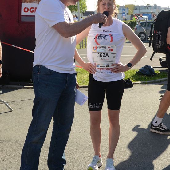 Ein Mann in einem weißen T-Shirt und Jeans interviewt eine Frau, die ein weißes Tanktop mit der Startnummer 362A trägt. Sie stehen auf einer Straße mit einem roten Container und einem Rucksack in der Nähe.