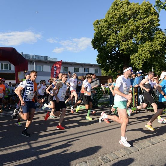 Eine Gruppe von Läufern bei einem Marathonrennen, mit einem Gebäude und einem Baum im Hintergrund.