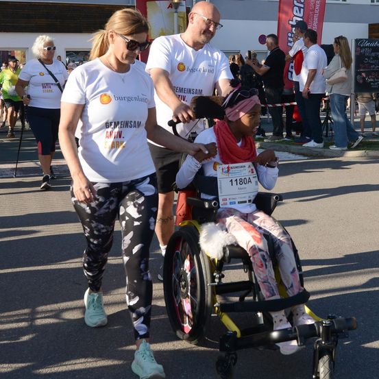 Ein junges Mädchen im Rollstuhl mit der Startnummer 1180A wird bei einem Marathon-Event von zwei Frauen geschoben.