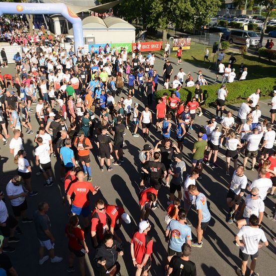 Eine große Menschenmenge, gekleidet in Laufausrüstung, versammelt sich am Start eines Marathons. Die Szene ist voller Aktivität, mit Teilnehmern in verschiedenen Farben und Größen von Shirts.