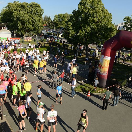 Eine große Menge von Läufern in verschiedenen farbigen Trikots versammelt sich am Start eines Marathons. Bäume und ein roter aufblasbarer Bogen sind im Hintergrund zu sehen.