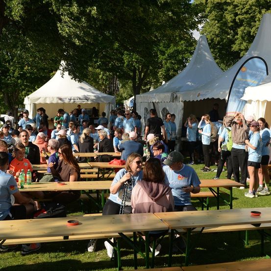 Eine Gruppe von Menschen in blauen Shirts versammelt sich in einem Außenbereich mit Zelten und Tischen. Einige sitzen, andere stehen. Der Bereich ist von Bäumen umgeben.