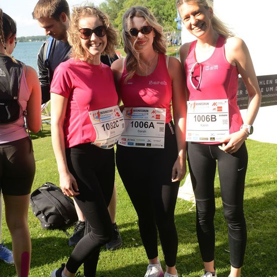 Drei Frauen in pinkfarbenen Shirts und schwarzen Leggings stehen auf einem Rasenfeld und lächeln für ein Foto. Sie halten Startnummern mit den Nummern 1082C, 1006A und 1006B. Im Hintergrund sind verschwommene Personen und ein See zu sehen.