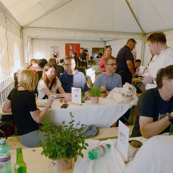 Eine Gruppe von Menschen sitzt an einem Tisch in einem Zelt, möglicherweise bei einer gesellschaftlichen Veranstaltung. Einige essen, während andere in Gespräche vertieft sind. Der Tisch ist mit Tellern, Gläsern und Topfpflanzen gedeckt. Ein Mann in einem weißen Hemd hält einen Becher.