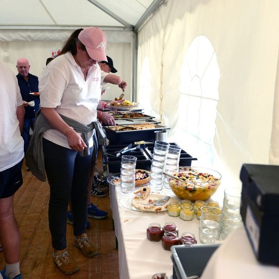 Eine Gruppe von Menschen steht in einer Schlange an einem Buffettisch mit verschiedenen Speisen und Getränken. Eine Frau mit einer rosa Mütze schaut sich das Essen an.