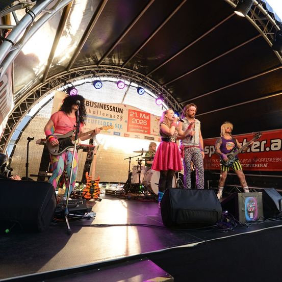 Eine Band tritt auf einer Bühne mit einem farbenfrohen Hintergrund auf, mit einem Schlagzeuger, Gitarristen und Sängern unter einem Scheinwerfer.