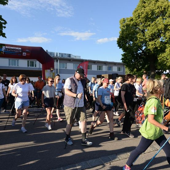 Eine Gruppe von Menschen nimmt an einer Nordic-Walking-Veranstaltung teil, trägt verschiedene Sportbekleidung und hält Wanderstöcke. Die Veranstaltung findet unter einem klaren blauen Himmel statt, mit einem Gebäude und Bäumen im Hintergrund.
