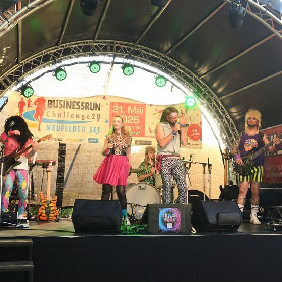 Eine Band tritt auf einer Bühne auf, hinter ihnen ein Banner mit der Aufschrift 'BUSINESSRUN Challenge2B'. Die Bandmitglieder tragen farbenfrohe Outfits und spielen verschiedene Instrumente.