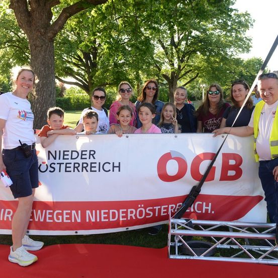 Eine Gruppe von Menschen, darunter Erwachsene und Kinder, posiert für ein Foto vor einem Banner mit der Aufschrift 'Nieder Osterreich'. Sie stehen auf einem roten Teppich.