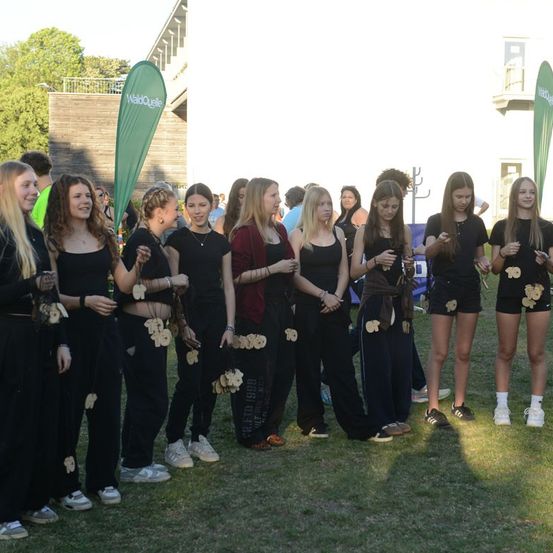 Eine Gruppe junger Frauen in schwarzen Kleidern mit Blumendesigns steht in einer Reihe auf einem Rasenfeld.