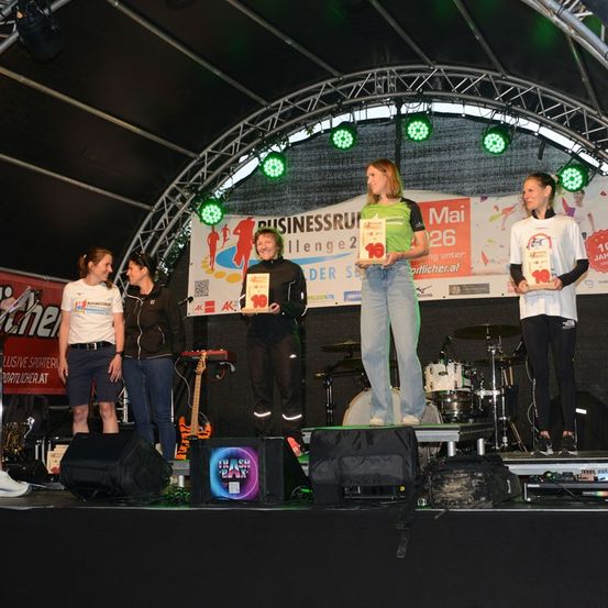 Eine Gruppe von Frauen steht mit Trophäen auf der Bühne, mit einem Banner im Hintergrund, das 'Mai 26' steht. Grüne Lichter beleuchten die Bühne.