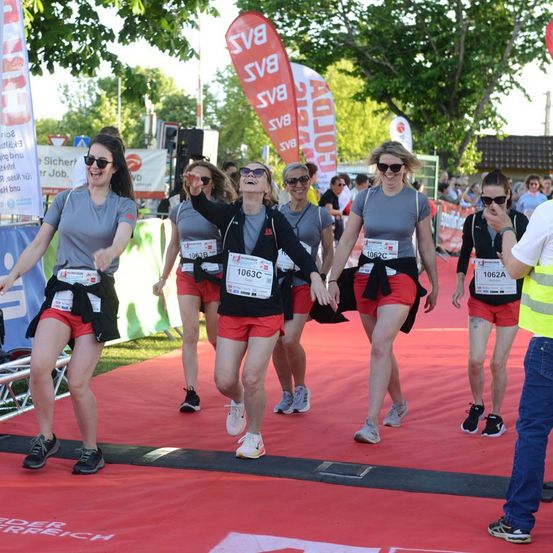 Eine Gruppe von Frauen in passenden grauen Oberteilen und roten Shorts läuft in einem Marathon. Sie lächeln und scheinen Spaß zu haben. Der Hintergrund zeigt einen roten Teppich, Banner und Bäume.
