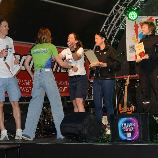 Eine Gruppe von Menschen steht auf einer Bühne mit einem Mikrofon, einem Pokal und einem Banner. Eine Frau hält ein Papier und eine andere Frau einen Pokal.