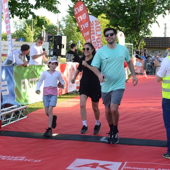 Eine Familie beendet einen Marathon auf einem roten Teppich, mit einem Mann in einem hellblauen Shirt, einer Frau in Schwarz und einem jungen Mädchen in Pink. Zuschauer und Banner sind im Hintergrund.