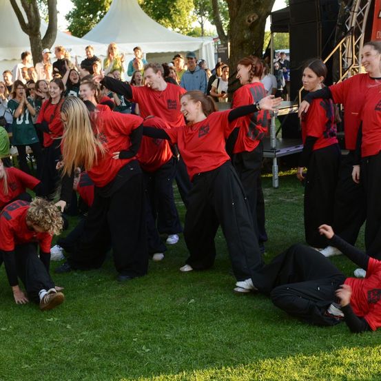 Eine Gruppe von Menschen in roten und schwarzen Outfits führt eine Tanzroutine auf einem Rasenfeld auf, während eine Menge zuschaut.