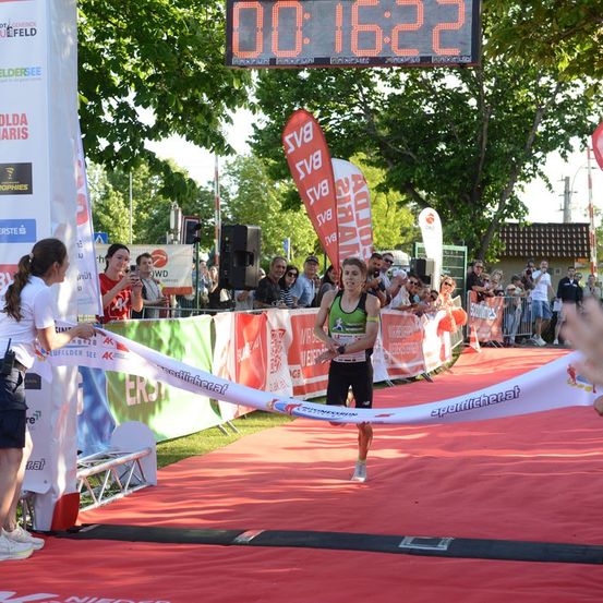 Ein Marathonläufer überquert die Ziellinie, wobei die digitale Uhr 00:16:22 anzeigt. Zuschauer beobachten hinter einem Zaun.
